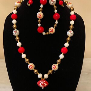 Könnte beinhalten: Ein geschmücktes Schmuckset mit Halskette, Armband und Ohrringen. Die Halskette hat rote, weiße und goldene Perlen mit einem Football-Anhänger. Die Ohrringe und das Armband passen zur Halskette, mit dem Kansas City Chiefs Logo.