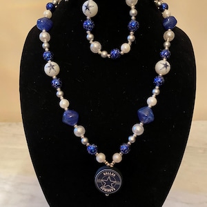 May include: A necklace, bracelet, and earrings set featuring blue, silver, and white beads. The necklace has a pendant with the Dallas Cowboys logo. The beads are various sizes and shapes, including round, square, and glittery.