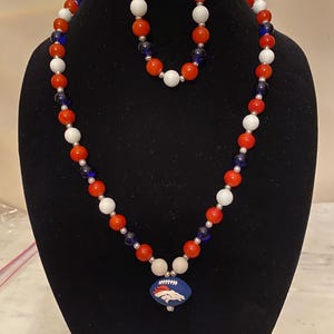May include: A necklace and bracelet set featuring red, white, and blue beads. The necklace has a pendant with a football design. The jewelry is displayed on a black mannequin.