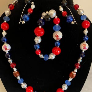 May include: A beaded jewelry set featuring a necklace, bracelet, and earrings. The necklace and bracelet have red, blue, silver, and white beads. The earrings have a black bead at the top. Some beads have a football design.