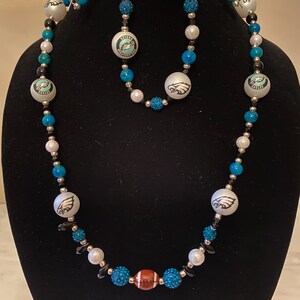 Può includere: Un set di collana e bracciale con perline bianche, blu e nere. La collana presenta grandi perline bianche con un logo di aquila, una perlina a forma di pallone da football e perline blu e nere più piccole. Il bracciale rispecchia il design della collana.