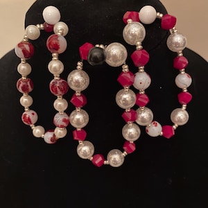 May include: A necklace with multiple strands of beads in red, white, and silver. The beads include large silver orbs, red faceted beads, small white pearls, and white beads with red splatter designs. The necklace is displayed on a black mannequin.