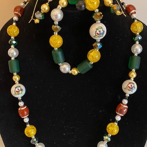 Może przedstawiać: Zestaw naszyjnika, bransoletki i kolczyków z różnymi koralikami. Naszyjnik i bransoletka zawierają żółte, zielone, srebrne i czerwone koraliki. Kolczyki są w kolorze złotym z czarnymi i białymi koralikami. Niektóre koraliki mają wzór piłki nożnej.