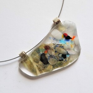 Op de afbeelding: Een ketting met een heldere, onregelmatig gevormde hanger en een zilverkleurige draad. De hanger is gevuld met kleurrijke stenen, waaronder rood, blauw en groen, en witte en heldere accenten. De ketting staat op een witte achtergrond.