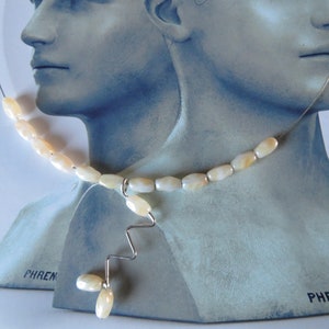 Può includere: Una collana con una base in filo sottile color argento decorata con perline lisce, ovali e color crema. Il design include un elemento centrale a goccia con un segmento di filo argentato a zigzag che termina con due perline pendenti.