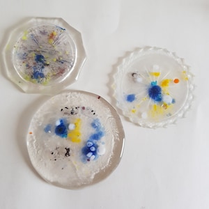 Op de afbeelding: Drie transparante glazen onderzetters met verschillende patronen van blauwe, gele en witte verfspatten.