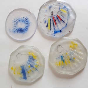 Può includere: Quattro pezzi decorativi in vetro con disegni astratti. I pezzi sono trasparenti con accenti colorati di blu, giallo e rosso. Uno è rotondo, mentre gli altri hanno forme irregolari. I disegni assomigliano a occhi stilizzati o motivi floreali.