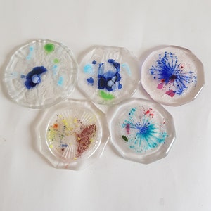 Op de afbeelding: Een set van vijf doorzichtige, achthoekige onderzetters met ingebedde kleurrijke inktpatronen. Elke onderzetter toont unieke abstracte ontwerpen in tinten blauw, groen, rood en geel, met sommige voorzien van stervormige effecten en andere met gespikkelde texturen.