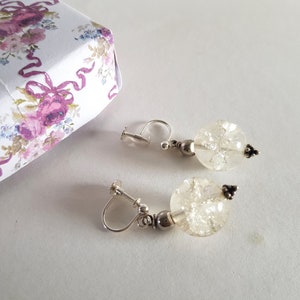 May include: A pair of clip-on earrings featuring clear, crackled glass beads. The earrings have silver-colored metal accents and a small, decorative black element. A floral patterned box is in the background.