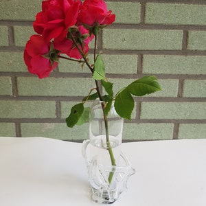 Könnte beinhalten: Eine klare Glasvase mit einem dekorativen, abstrakten, skulpturalen Rahmen, der einen einzelnen Stängel mit drei leuchtend roten Rosen und grünen Blättern hält. Die Vase ist mit Wasser gefüllt und hat einen markanten, transparenten Glasfuß.