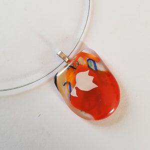 Op de afbeelding: Een kleurrijke hangerketting met een zilverkleurige draad. De hanger heeft een heldere, onregelmatige vorm met abstracte patronen in rood, oranje, blauw en wit. De hanger is met een zilverkleurige sluiting aan de draad bevestigd.