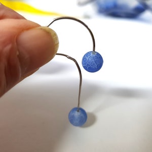 Op de afbeelding: Zilverkleurige oorbellen met een gebogen ontwerp. Elke oorbel heeft een kleine, ronde, blauwe kraal met een gebarsten, getextureerd oppervlak. De kralen zijn opgehangen aan het gebogen metaal.