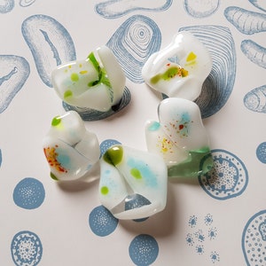Op de afbeelding: Vijf onregelmatige, doorschijnende witte glazen stukken met verspreide insluitingen van groene, blauwe, gele en oranje spikkels. Elk stuk heeft een gladde, afgeronde en organische vorm met variërende interne kleurpatronen en heldere delen.