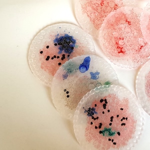 May include: A collection of circular, translucent coasters with a textured, scalloped edge. Each coaster features a unique abstract design with splashes of pink, blue, and black ink, creating a layered, organic appearance on the semi-clear surface.