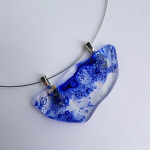 Puede incluir: Un collar con un colgante de vidrio azul y blanco con una cadena de plata. El colgante tiene una forma única e irregular y presenta pequeñas burbujas azules en todo el vidrio.