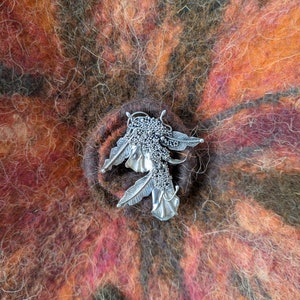 Può includere: Una spilla color argento caratterizzata da un gruppo di sfere granulose testurizzate e forme simili a foglie, fissata su una superficie in lana infeltrita con un motivo variegato di fibre marrone scuro, arancione bruciato e rosso vivo.