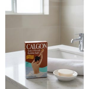 Op de afbeelding: Een doos Calgon Spice Woods Bubble Bath met een bruin en blauw ontwerp. De doos bevat een illustratie van een persoon in een bubbelbad. Een witte zeep is in een klein wit schaaltje.