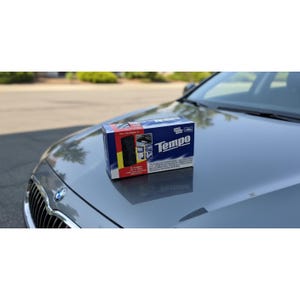 Peut inclure: Une boîte bleue, rouge et blanche avec le logo "Tempo" est posée sur le capot d'une voiture grise. La boîte contient des images et du texte, suggérant un étui pour voiture. La voiture est garée à l'extérieur par une journée ensoleillée.