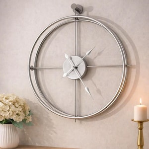 May include: A large, silver-colored wall clock with a minimalist design. The clock features a circular frame with a smaller central dial and slender hands. The clock is mounted on a light-colored wall, with a vase of flowers and a candle holder nearby.