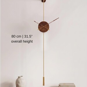 May include: A modern wall clock with a wooden face and brass accents. The clock has a pendulum and measures 31.5 inches in overall height. The clock is mounted on a white wall, with a shelf and vase below.