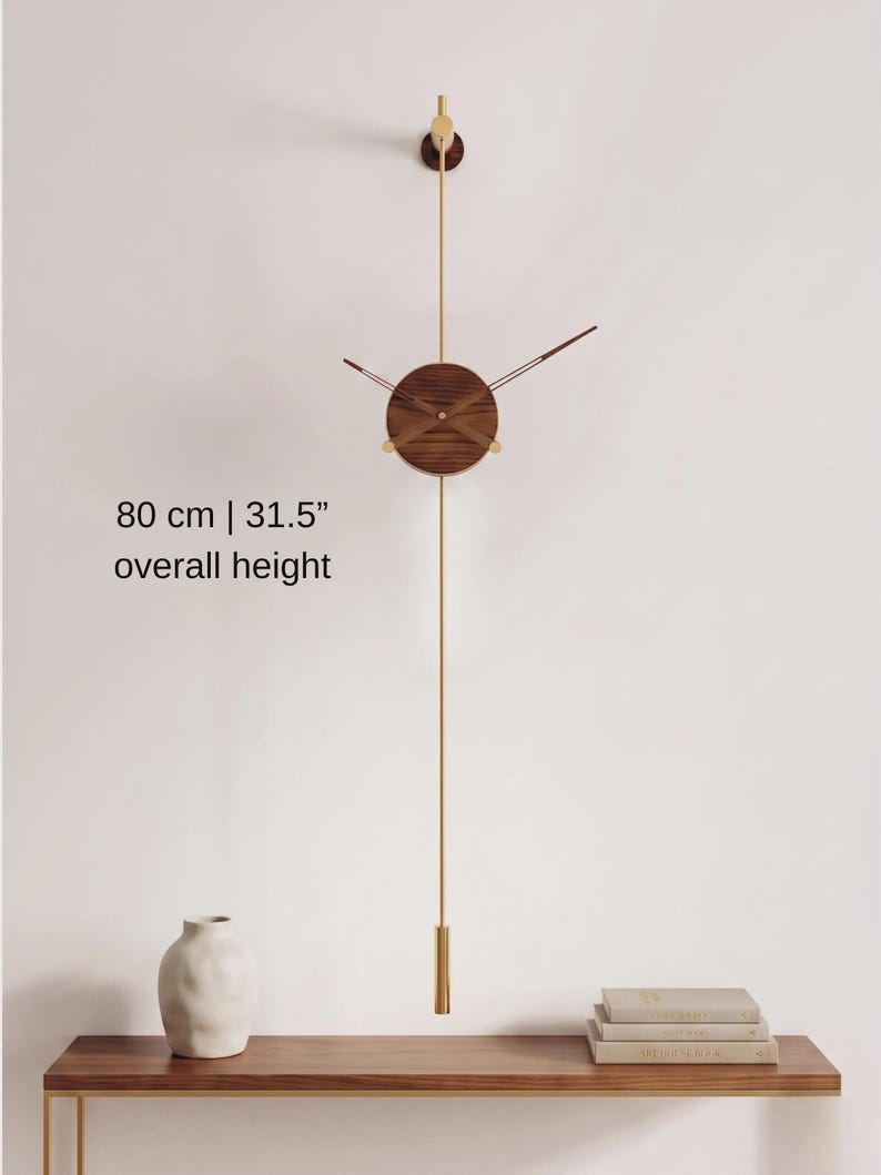 May include: A modern wall clock with a wooden face and brass accents. The clock has a pendulum and measures 31.5 inches in overall height. The clock is mounted on a white wall, with a shelf and vase below.