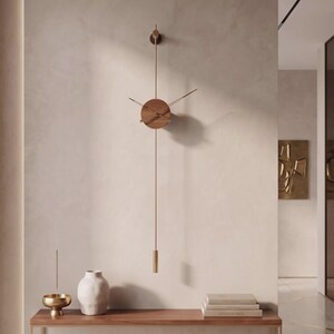 May include: A modern wall clock with a round wooden face and brass hands and pendulum. The clock is mounted on a light-colored wall above a wooden console table. A white vase, a gold bowl, and books are on the table.