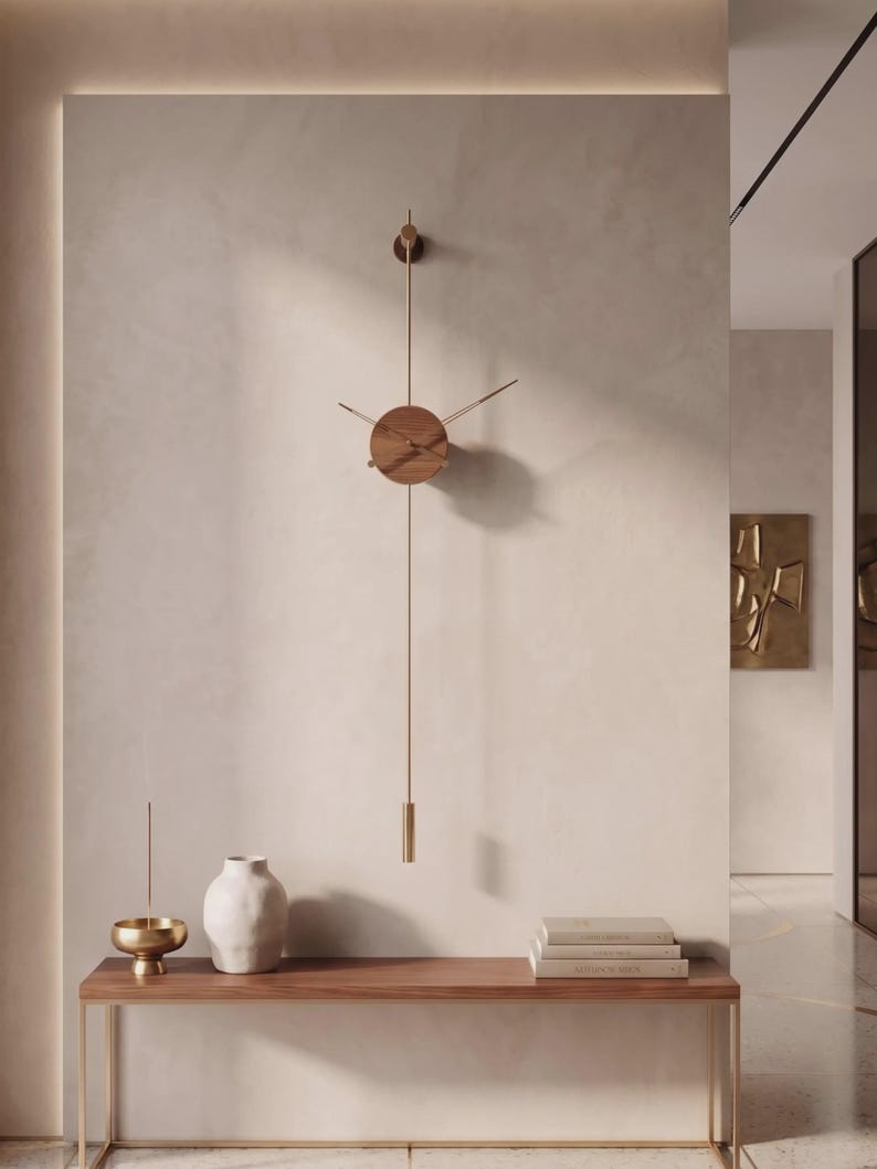 May include: A modern wall clock with a round wooden face and brass hands and pendulum. The clock is mounted on a light-colored wall above a wooden console table. A white vase, a gold bowl, and books are on the table.