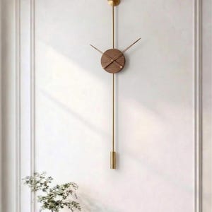 May include: A modern wall clock with a round, wooden face and gold-colored hands. The clock hangs from a gold-colored rod with a decorative element at the top and bottom. The clock is mounted on a white wall.
