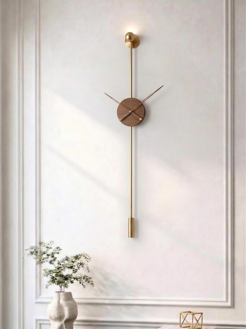 May include: A modern wall clock with a round, wooden face and gold-colored hands. The clock hangs from a gold-colored rod with a decorative element at the top and bottom. The clock is mounted on a white wall.
