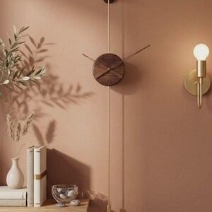 May include: A modern wall clock with a round, dark wood face and gold-colored hands and pendulum. The clock is mounted on a muted orange wall, with a gold sconce and a small shelf with books and decorative items.