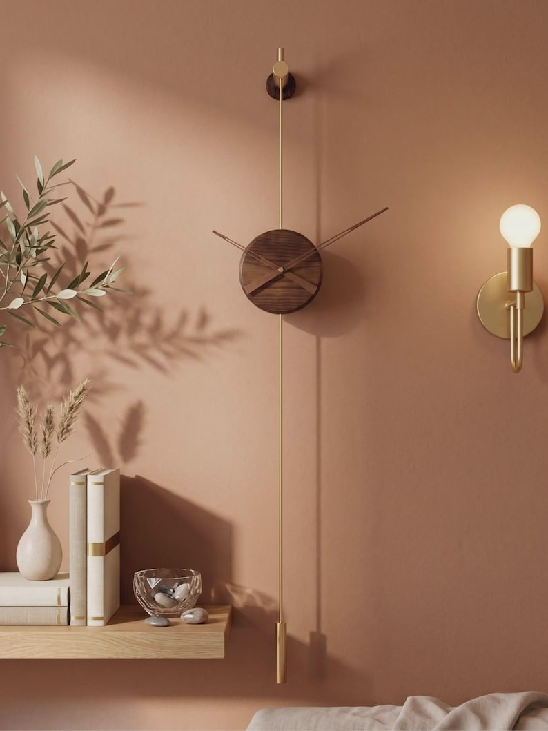 May include: A modern wall clock with a round, dark wood face and gold-colored hands and pendulum. The clock is mounted on a muted orange wall, with a gold sconce and a small shelf with books and decorative items.