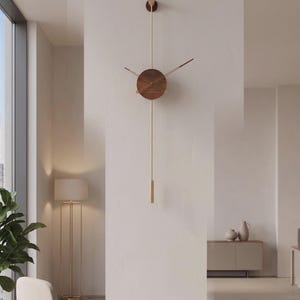 May include: A modern wall clock with a minimalist design. The clock features a round, wooden face with brass-colored hands and a long, brass-colored pendulum. The clock is mounted on a white wall in a well-lit room.