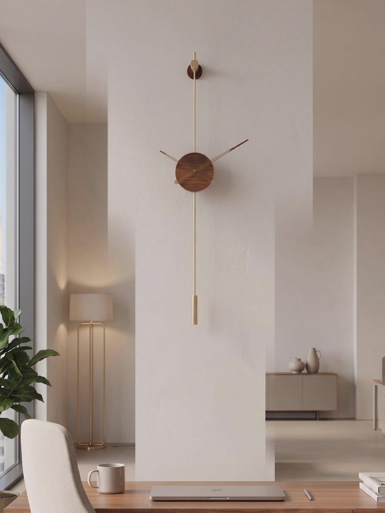May include: A modern wall clock with a minimalist design. The clock features a round, wooden face with brass-colored hands and a long, brass-colored pendulum. The clock is mounted on a white wall in a well-lit room.