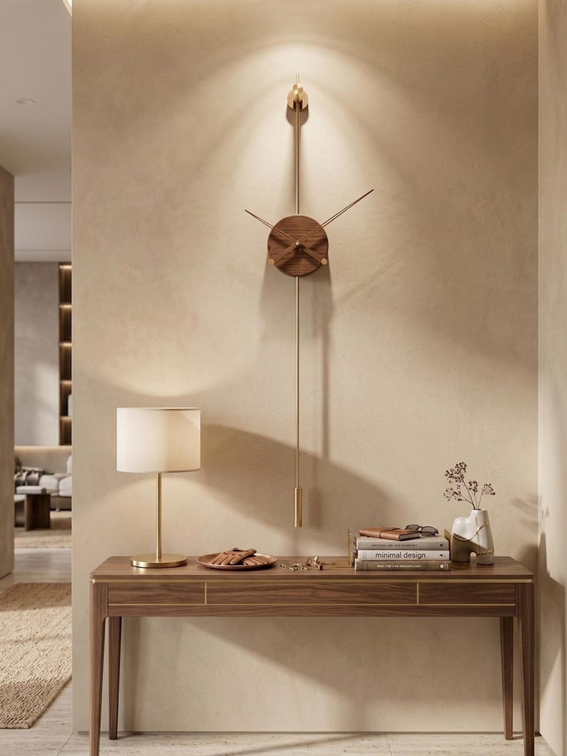 May include: A modern wall clock with a wooden face and gold-colored accents. The clock has a long pendulum and is mounted on a beige wall. A wooden console table with a lamp and decorative items is below the clock.