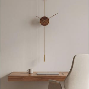 May include: A modern wall clock with a round, wooden face and brass hands and pendulum. The clock hangs on a light-colored wall above a wooden desk with a laptop and mug. The clock's design is minimalist and elegant.