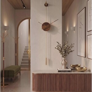 May include: A modern, minimalist wall clock with a round, wooden face and gold-tone hands and pendulum. The clock is mounted on a light-colored wall, and the image showcases a hallway with a bench, artwork, and a console table.