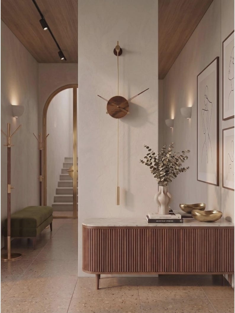 May include: A modern, minimalist wall clock with a round, wooden face and gold-tone hands and pendulum. The clock is mounted on a light-colored wall, and the image showcases a hallway with a bench, artwork, and a console table.
