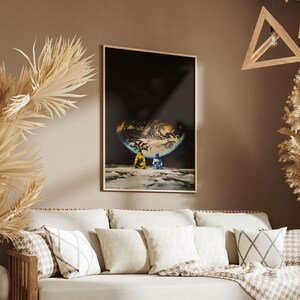 May include: A rectangular wall art print in a thin wooden frame featuring a dark space scene with a glowing, partially illuminated planet surface. Two small figures in yellow and blue clothing sit on a cratered lunar landscape looking at Earth.