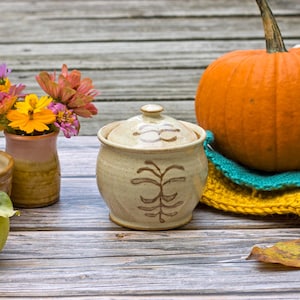 Pode incluir: Natureza morta com uma abóbora laranja, um pequeno pote de cerâmica com tampa, uma tigela, um vaso de flores e uma maçã verde. Uma pinha e um item de crochê amarelo e turquesa também estão presentes. Em uma superfície de madeira envelhecida.