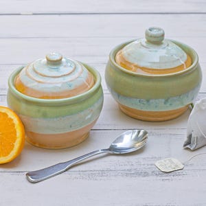 Pode incluir: Dois potes de cerâmica com tampas listradas em laranja, verde e azul, uma colher de prata, um saquinho de chá e uma fatia de laranja. Os potes são redondos com um pequeno botão no topo. Uma flor rosa com centro amarelo está ao lado do saquinho de chá.