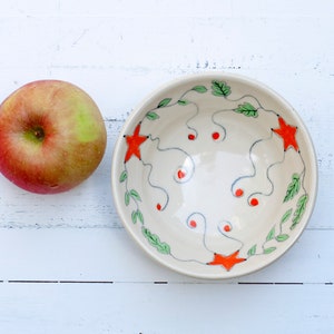 May include: A white ceramic bowl with a hand-painted design of green leaves, orange stars, and red dots. The bowl is on a white wooden surface next to a red apple.
