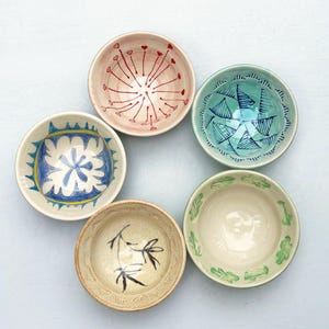 May include: Five ceramic bowls arranged in a circle. One bowl is pink with red line patterns, one is light blue with dark blue triangles, one is white with blue and yellow designs, one is beige with black brush strokes, and one is cream with green leaves.