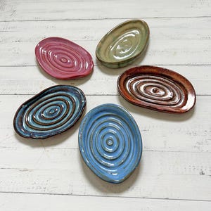 May include: Five handmade ceramic oval dishes in shades of red, green, brown, blue, and teal. Each dish features a concentric spiral design in the center. The dishes are arranged on a white wooden surface.
