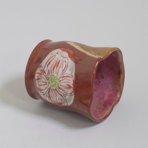 May include: A handmade ceramic mug with a floral design. The exterior is a reddish-brown color, while the interior is a vibrant pink. A white flower with red and green accents is painted on the side. The mug has a unique, organic shape.