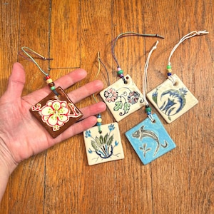 May include: Five square ceramic ornaments with hand-painted designs including a red flower, floral patterns, a blue fish, and green plants. Each ornament features a hole at the top with a string and small colorful beads for hanging.