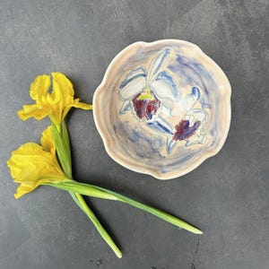 May include: A ceramic bowl with a scalloped edge, featuring a hand-painted floral design in white, blue, and burgundy. Two yellow iris flowers with green stems are placed beside the bowl on a dark gray surface.