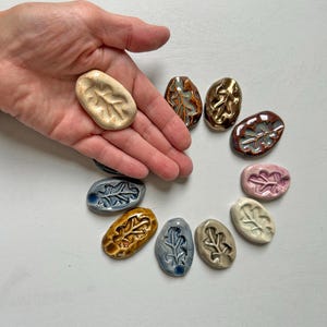 Peut inclure: Une collection de onze pierres en céramique ovales, chacune présentant un motif de feuille en relief. Les pierres sont finies avec divers émaux brillants et mats, incluant des tons crème, or métallique, brun, rose, bleu et gris.