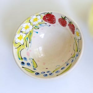 May include: A ceramic bowl with a cream-colored interior and a hand-painted floral and fruit design. The bowl features white flowers with yellow centers, red strawberries, and blue blueberries. Green and yellow accents complete the design.