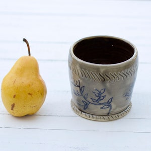 May include: A gray ceramic mug with a blue floral design. The mug has a textured rim and a slightly flared lip. The mug is sitting on a white surface.
