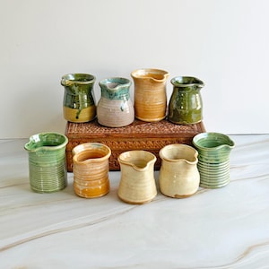 May include: A selection of handmade ceramic vases in shades of green, tan, and brown. The vases have an organic form with a small spout. Some are striped, others smooth. They are displayed on a wooden box and a marble surface. Measurements are in cm.